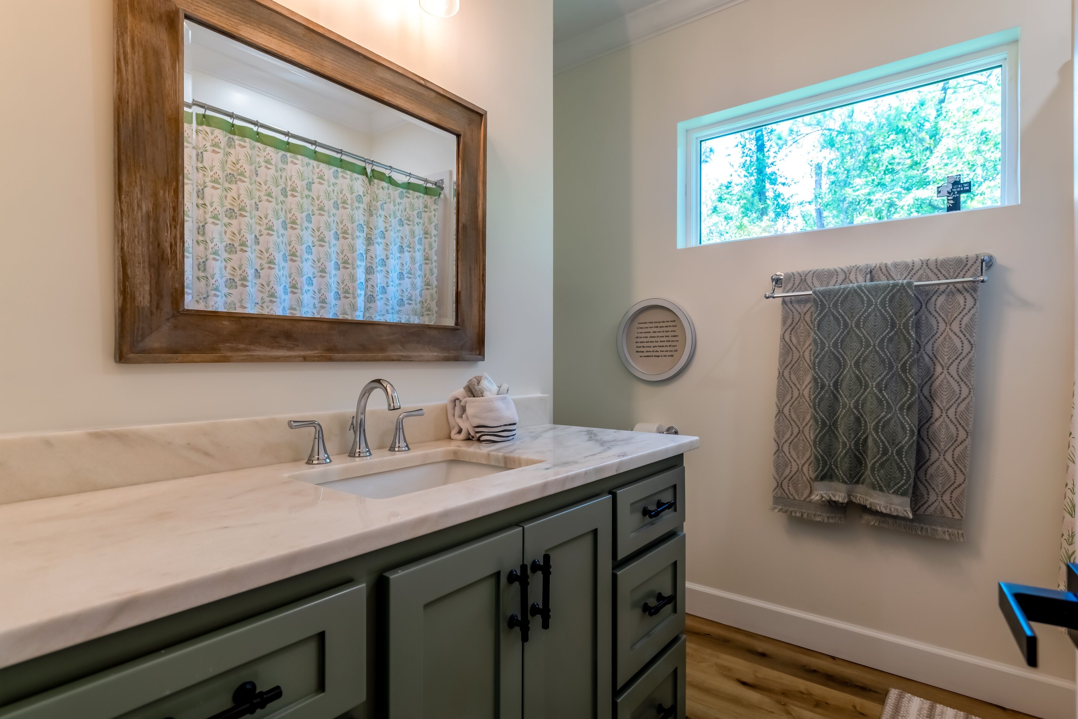 A bathroom with a marble sink, green cabinets, a wooden-framed mirror, a window with a view of trees, and patterned towels.