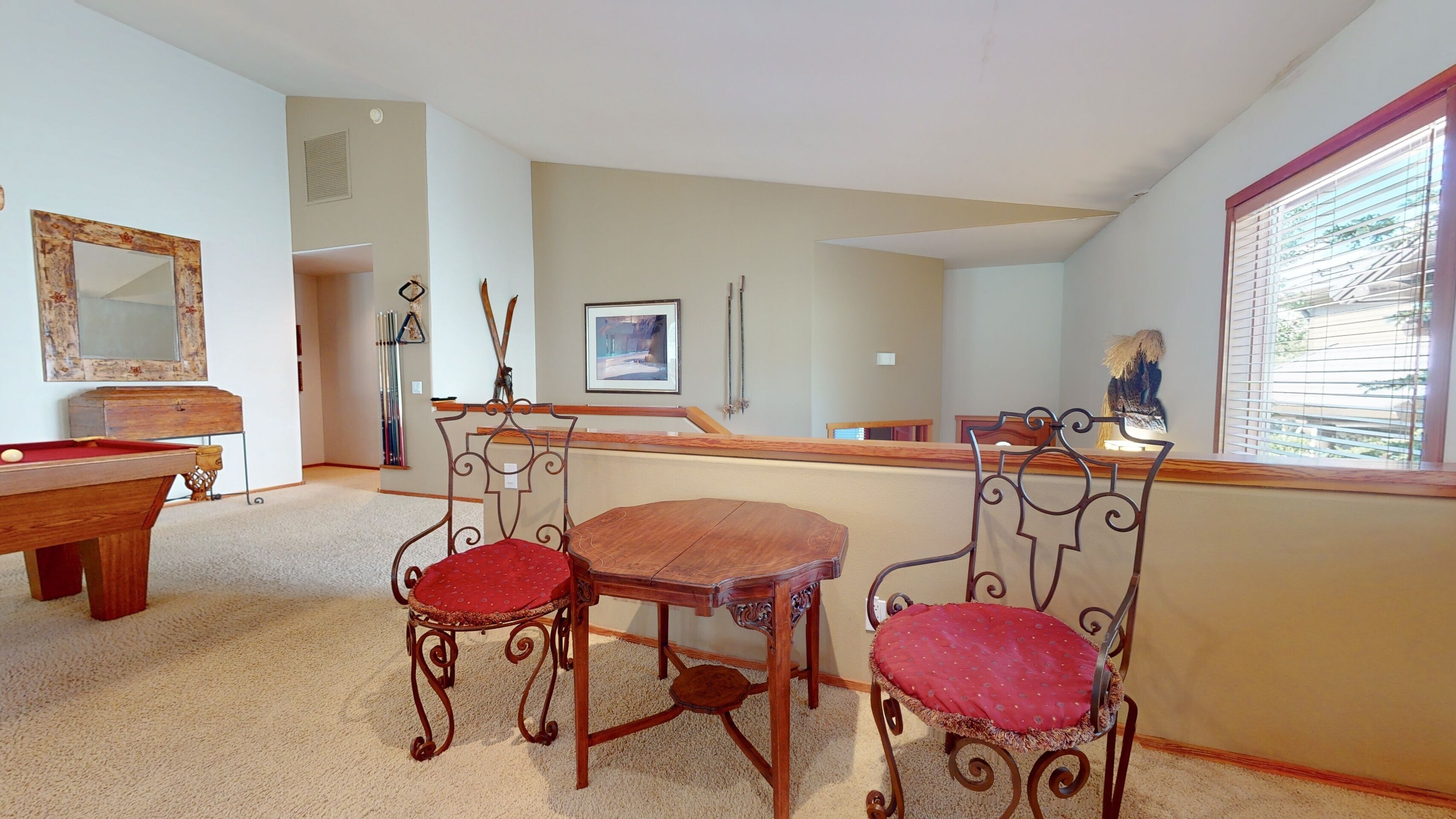 A room with a pool table, two ornate chairs with red cushions, a wooden table, and a window with blinds.
