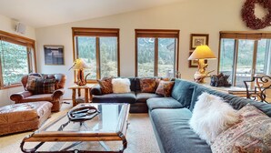 A cozy living room with a leather sofa, a glass coffee table, and a fireplace. There are large windows with a view of trees and mountains.