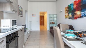 A modern kitchen with a microwave, oven, and cabinets. There is a dining area with a table set for two, a sofa, and a glass cabinet. A colorful painting is on the wall. - Manzoni Apartment (Roma)