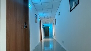 Un pasillo largo y estrecho con una puerta de madera a la izquierda, un cuadro enmarcado a la derecha y una señal de salida iluminada en azul al final.