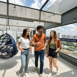 Three people standing on a balcony with a hanging chair, large windows, and a city view. - Audaar Tech Suítes (São Paulo)