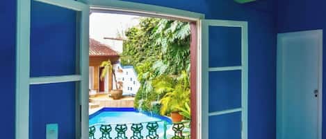 An open door leading to a balcony with a tiled floor, a pool, and a view of a house and greenery.