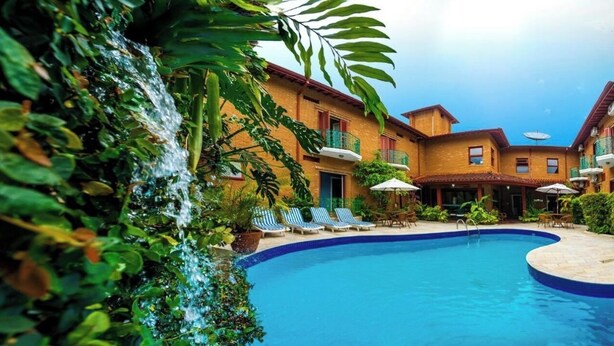 Piscina com espreguiçadeiras e guarda-sóis, rodeada por vegetação exuberante e um edifício com varandas.
