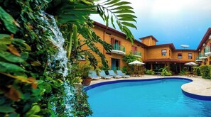 Piscina com espreguiçadeiras e guarda-sóis, rodeada por vegetação exuberante e um edifício com varandas.