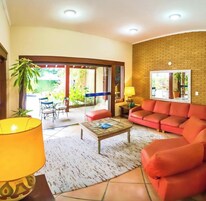Una sala de estar con un sofá, una mesa de centro y una lámpara. Hay un ventanal que da a un área exterior con sillas y una mesa.