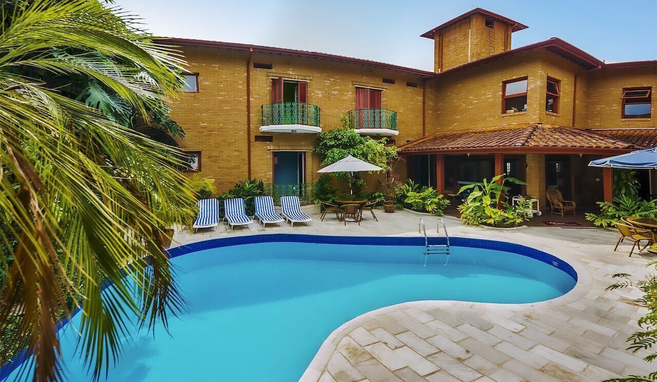 A swimming pool with lounge chairs and umbrellas, surrounded by a building with balconies and a tiled roof.