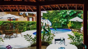 Um pátio externo com uma mesa redonda e cadeiras, cercado por vegetação exuberante e uma piscina.