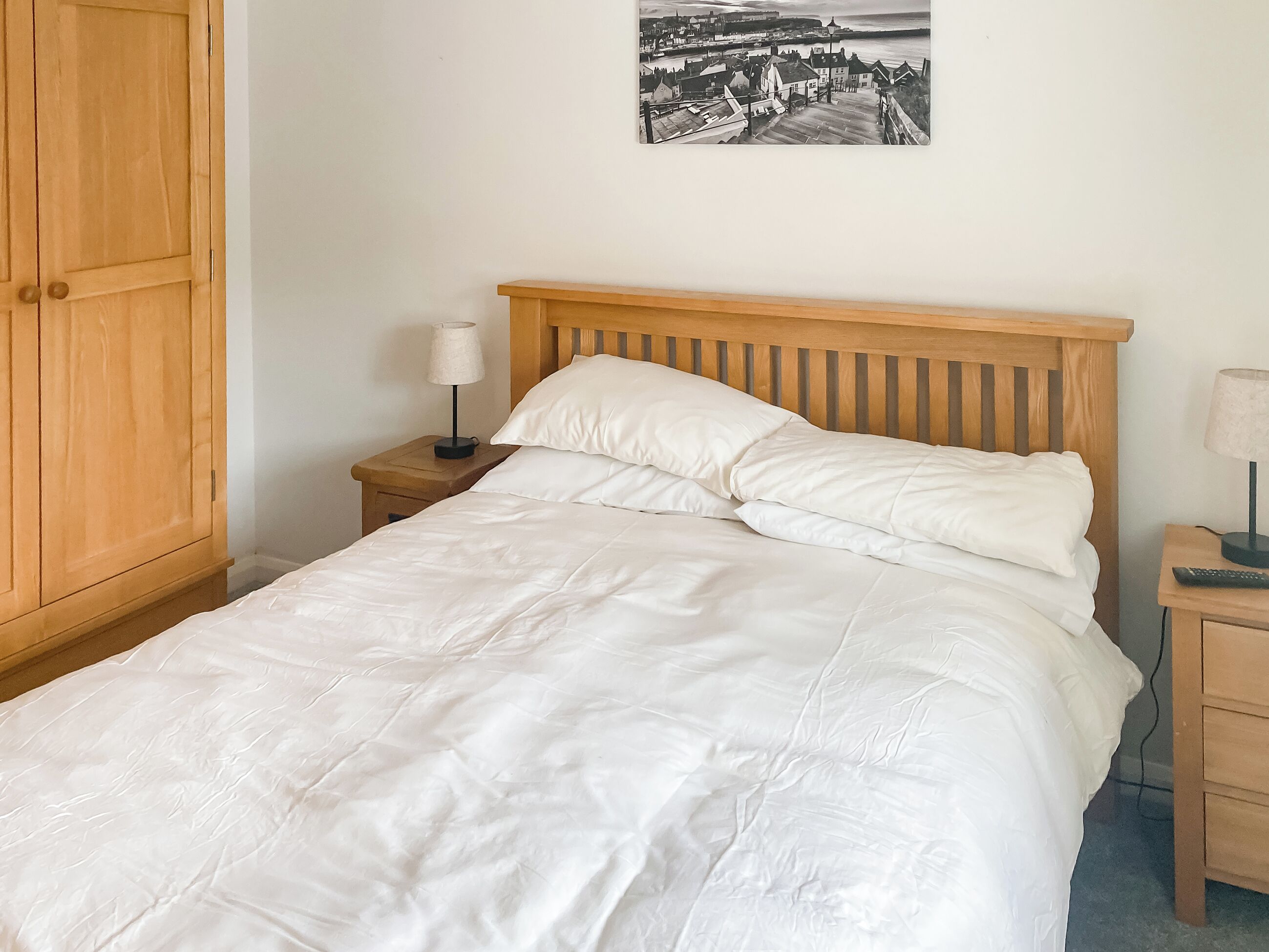 A bedroom with a wooden bed, white bedding, a nightstand with a lamp, a wooden wardrobe, and a black and white photo on the wall.
