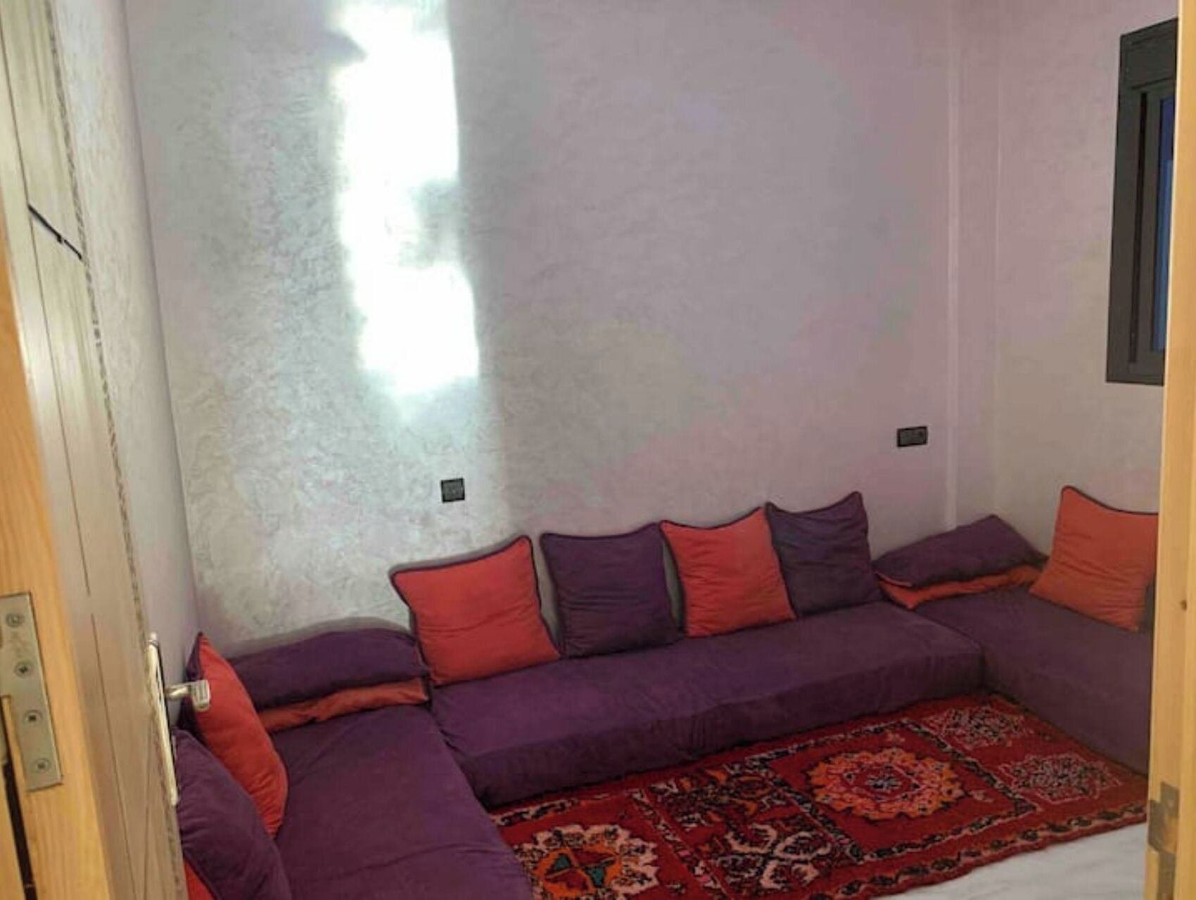 A room with a purple sofa, red and purple cushions, and a patterned rug.