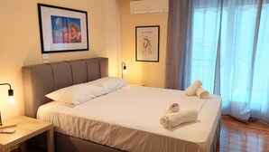 A neatly made bed with white linens and two rolled towels, a framed artwork above the bed, and a bedside table with a lamp. - Grand apartment in Acropolis museum (Athina)