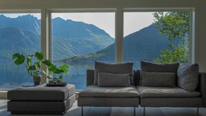 A modern living room with a grey sofa and ottoman, a potted plant, and a view of mountains and a lake through large windows. - Polar Panorama Lodge (Senja)