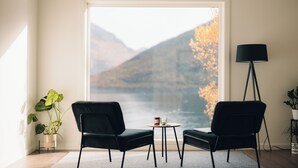 A room with two black chairs, a small round table with a candle and glass, a floor lamp, and a large window with a view of mountains and autumn foliage. - Polar Panorama Lodge (Senja)