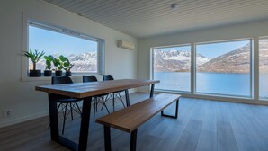 A modern dining area with a wooden table and benches, large windows offering a view of a lake and mountains, and potted plants on the windowsill. - Polar Panorama Lodge (Senja)