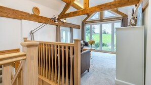Ein geräumiges Zimmer mit hoher Decke und freiliegenden Holzbalken, ein großes Fenster mit Blick auf eine grüne Landschaft, eine gemütliche Sitzecke mit Sofa und Tisch sowie eine moderne Lampe.