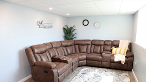 A brown leather sectional sofa with a throw blanket and pillows, a patterned rug, a wall clock, and a decorative wall piece.