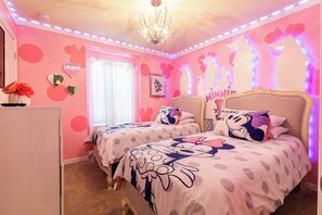 Een roze slaapkamer met twee bedden, elk met beddengoed van Minnie Mouse, een kroonluchter en een raam met gordijnen.
