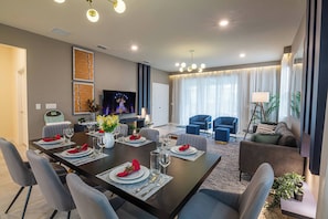 A modern living room with a dining area, featuring a dark wooden table set for six, grey upholstered chairs, a large flat-screen TV, and a comfortable seating area with a sofa and armchairs. - Beautiful Home with private Pool - SPA - Game Room (Davenport)