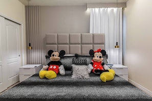 A bedroom with a grey upholstered headboard, a black and grey speckled bedspread, two Mickey Mouse plush toys, a white bedside table, and a white nightstand. - Beautiful Home with private Pool - SPA - Game Room (Davenport)