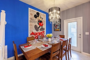 A dining room with a wooden table set for four, a Mickey Mouse poster, and a chandelier.