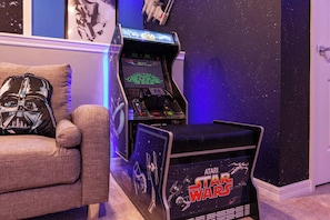 A Star Wars themed arcade with a console and a cabinet, a couch with a Darth Vader pillow, and a starry wall.