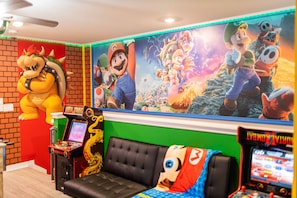 Quarto com um mural grande de Mario, um sofá com almofada do Mario e jogos de fliperama.