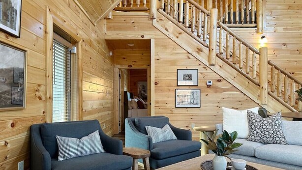 A wooden cabin interior with a staircase, two sofas, a coffee table, and framed pictures on the walls. - Luxury Smoky Mountain Cabin w/ Pool, Arcade, Hot Tub & Scenic Deck (Pigeon Forge)