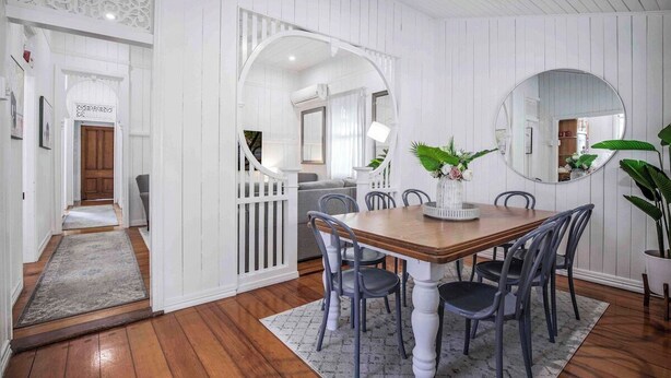 A dining area with a wooden table and chairs, surrounded by white paneled walls and a large mirror.