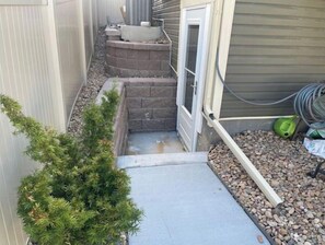 A narrow outdoor pathway leading to a door, flanked by a small tree and a stone planter.