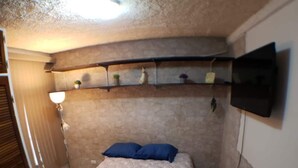 Un dormitorio pequeño y tenuemente iluminado con una cama individual, un estante con objetos decorativos y un televisor fijado a la pared.