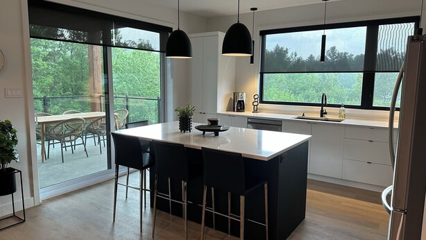 A modern kitchen with a central island, black pendant lights, and a large window offering a view of greenery. - The JOFORD, Condo, 5 minutes from skiing, Sépaq with extras (bikes, paddles +) (Orford)