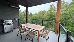 A covered outdoor dining area with a table and chairs, a grill, and a view of the forest. - The JOFORD, Condo, 5 minutes from skiing, Sépaq with extras (bikes, paddles +) (Orford)