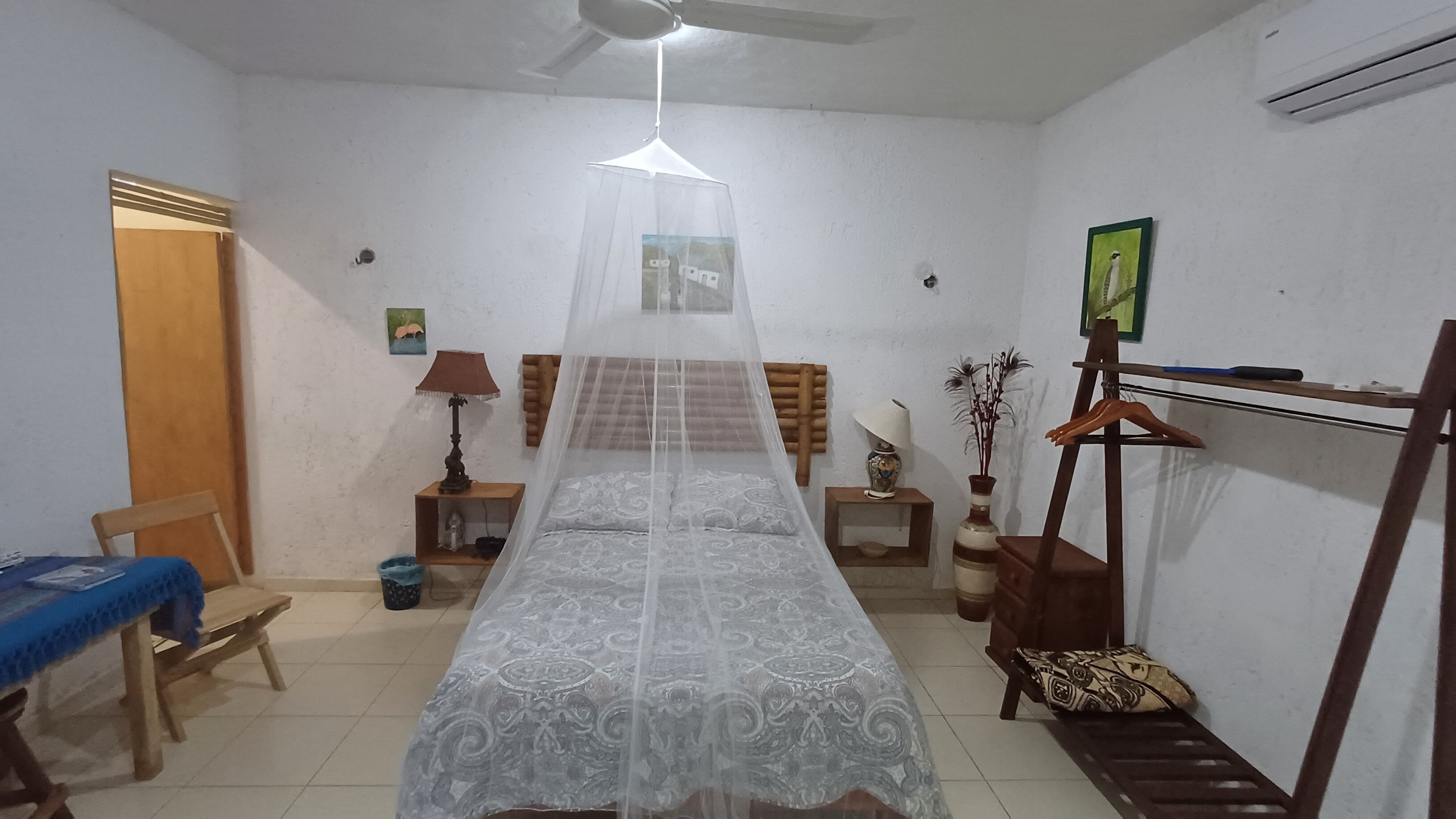 Un dormitorio con una cama cubierta por un dosel ligero, un cabecero de madera, una mesita de noche con una lámpara, una pequeña mesa con un florero y una silla.