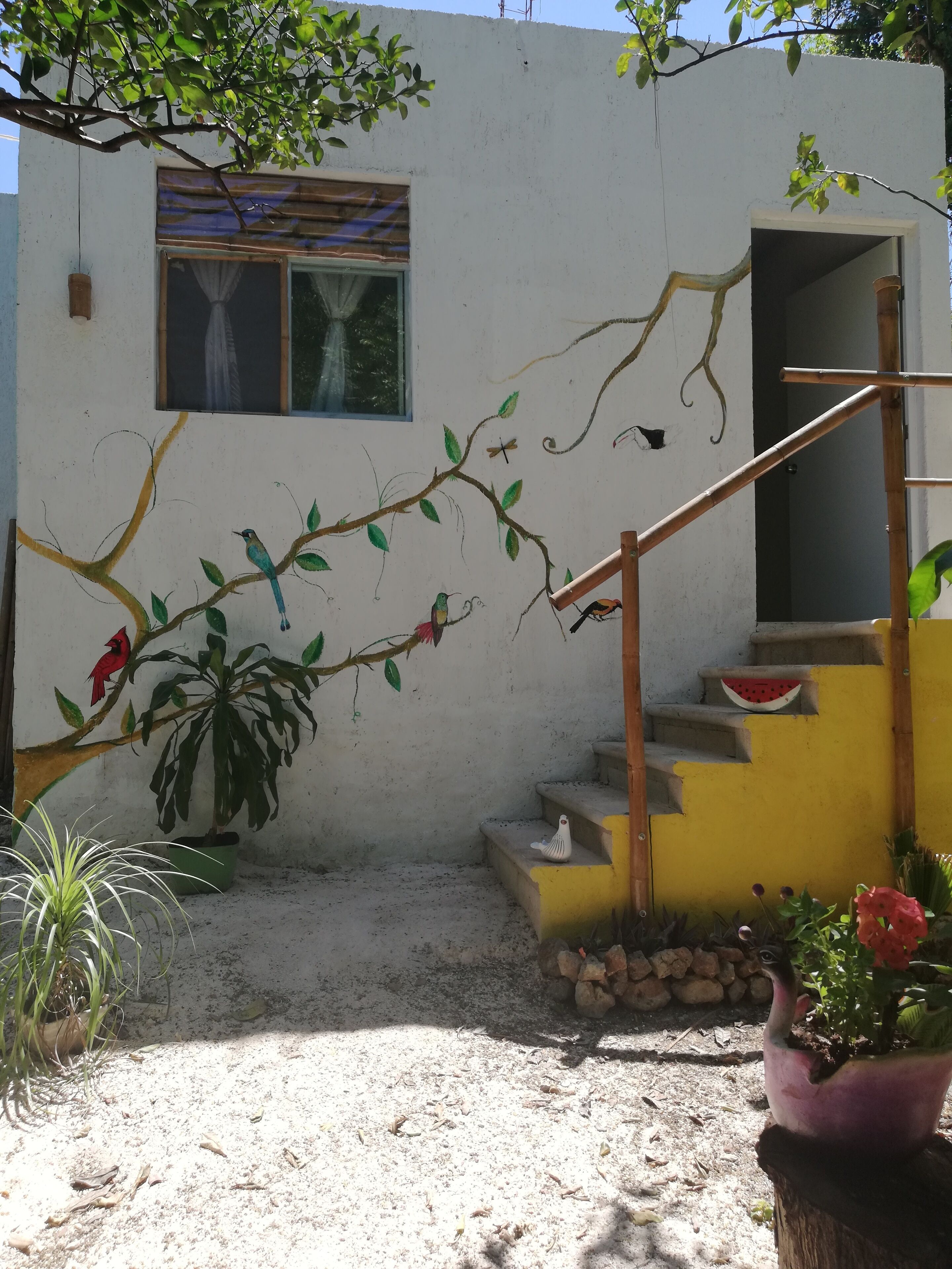 Una casa con una escalera que lleva a una puerta, una ventana con cortina, y una rama con hojas y pájaros pintados en la pared.