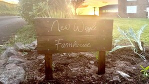 A wooden sign with "New Widge Farmhouse" written on it, set against a sunset background with a building and a road. - Rame View - Double Bedroom with En-suite (Millbrook)