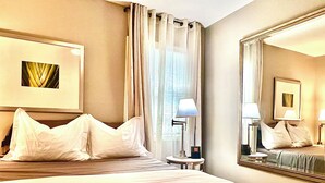 Habitación de hotel con una cama grande, una mesita de noche con lámpara, un espejo y una ventana con cortinas.