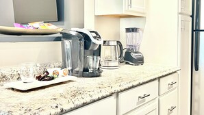 Una cocina con encimera de granito, cafetera, licuadora y hervidor. Hay armarios blancos y un refrigerador de acero inoxidable.