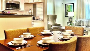 Un comedor con mesa dispuesta para seis personas, una cocina con muebles blancos y un microondas, y una sala de estar con un sillón grande y una planta.