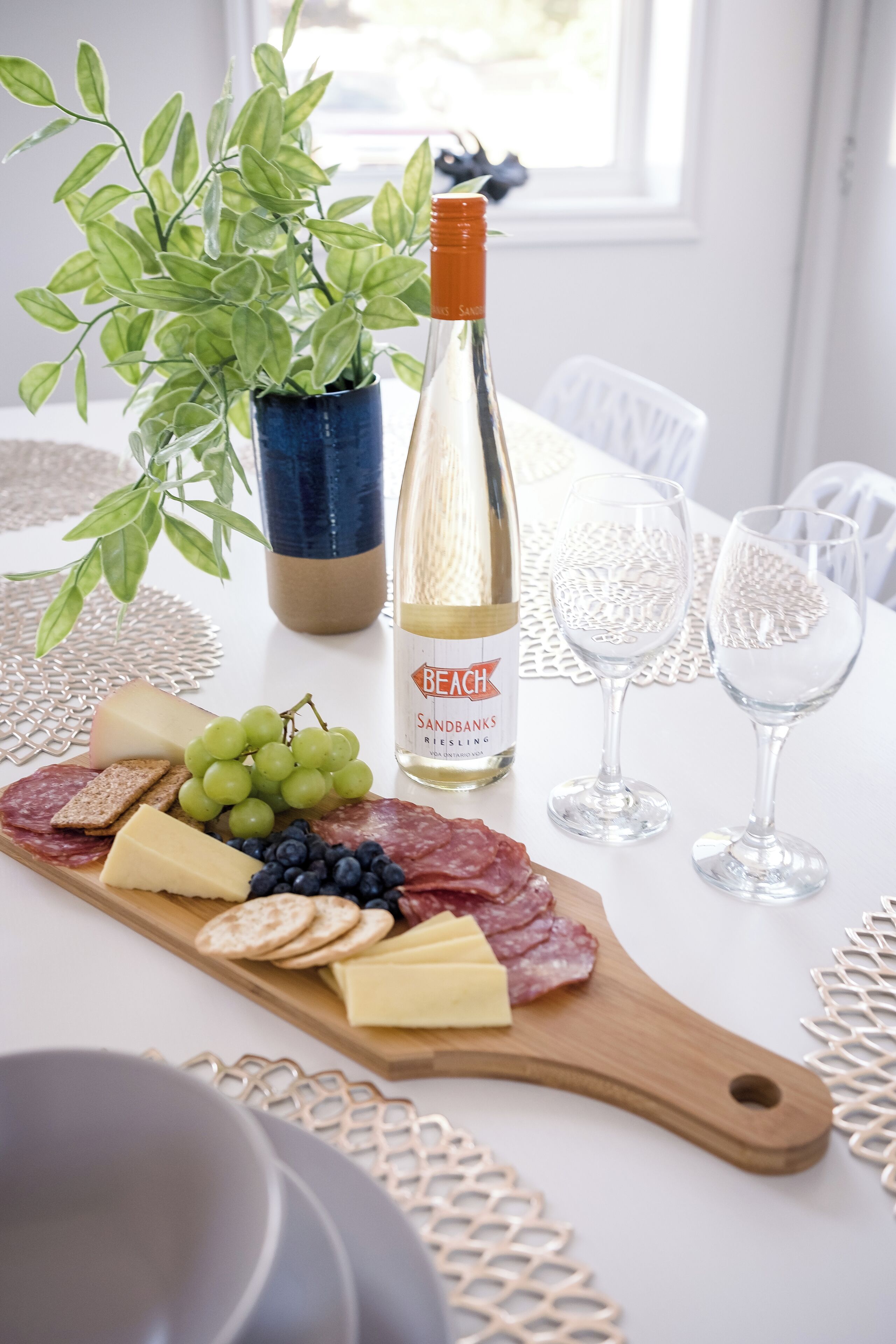 A table with a wooden board of cheeses, grapes, and crackers, a bottle of wine, and two glasses.