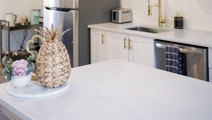 A modern kitchen with white marble countertops, a stainless steel refrigerator, a gold faucet, and geometric pendant lights.
