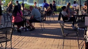 An outdoor dining area with tables and chairs, red umbrellas, and a view of a lake and trees.