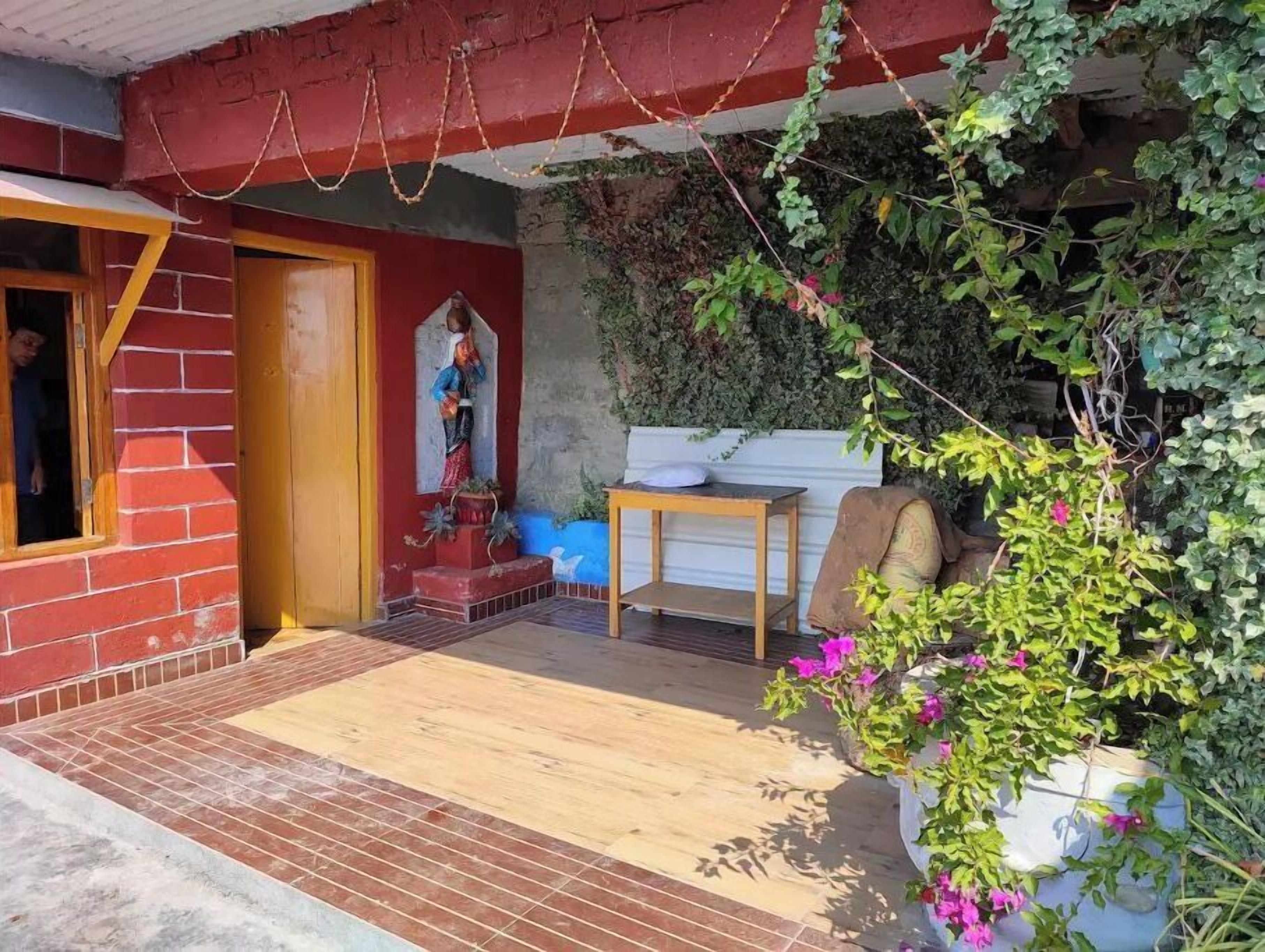 A small, rustic building with a red brick exterior, a wooden door, and a small porch area with a bench and a table.