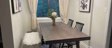 Un comedor con una mesa de madera, seis sillas, un banco, un jarrón con flores y una ventana con cortinas.