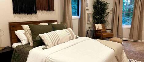 A bedroom with a large bed, a nightstand, a chair, and a potted plant.