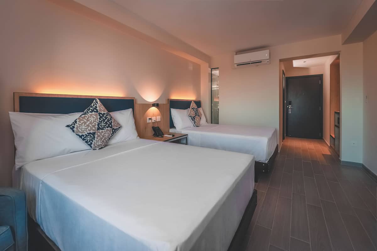 Une chambre d’hôtel avec deux lits, chacun doté d’une tête de lit, de tables de chevet et de lampes. Une porte donne accès à une autre pièce, et un appareil de climatisation est fixé au mur.