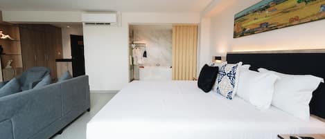 A hotel room with a large bed, a grey sofa, a bathroom with a marble wall, and a painting on the wall.