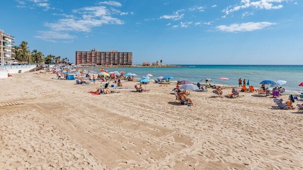 Ein Strand mit vielen Sonnenschirmen und Liegestühlen, ein großes Hotel im Hintergrund und strahlend blauer Himmel.