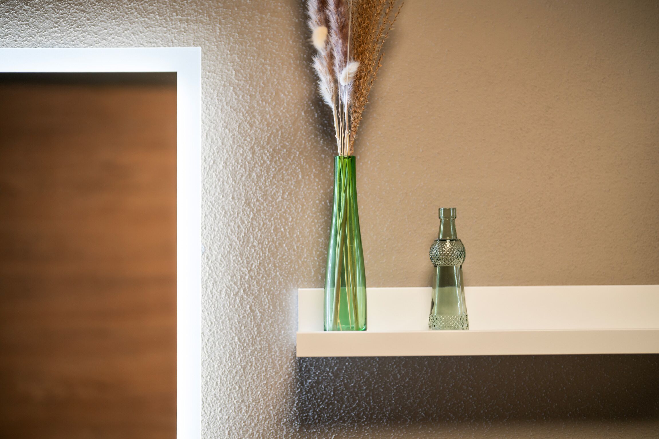 A green vase with dried plants on a white shelf against a textured wall.