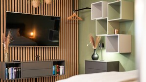 A modern bedroom with a TV mounted on a wooden panel wall, a bedside table with a lamp, and a shelf with books.