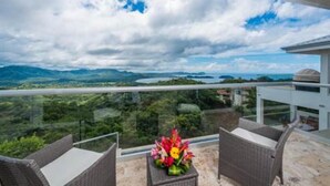 Ein Balkon mit Korbstühlen und Blumen, mit Blick auf eine Berglandschaft und ein Gewässer.